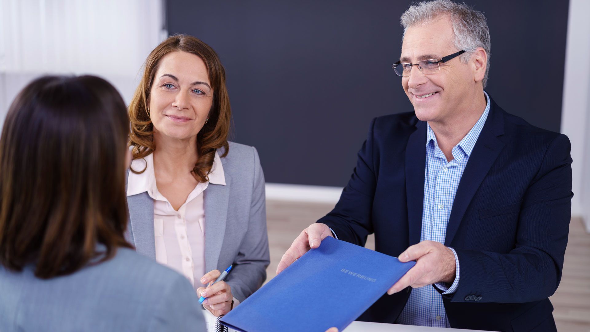 Zwei Fachpersonen überreichen in einem Meeting einen blauen Dokumentenordner an eine Kundin im Vordergrund. Die Szene vermittelt strukturierte Zusammenarbeit, Verlässlichkeit und Compliance—ideal zur Illustration unabhängiger Prüfung, Auditvorbereitung und Zertifizierungsprozesse in regulierten, sicherheitskritischen Branchen. Zwei Fachpersonen überreichen in einem Meeting einen blauen Dokumentenordner an eine Kundin im Vordergrund. Die Szene vermittelt strukturierte Zusammenarbeit, Verlässlichkeit und Compliance—ideal zur Illustration unabhängiger Prüfung, Auditvorbereitung und Zertifizierungsprozesse in regulierten, sicherheitskritischen Branchen.