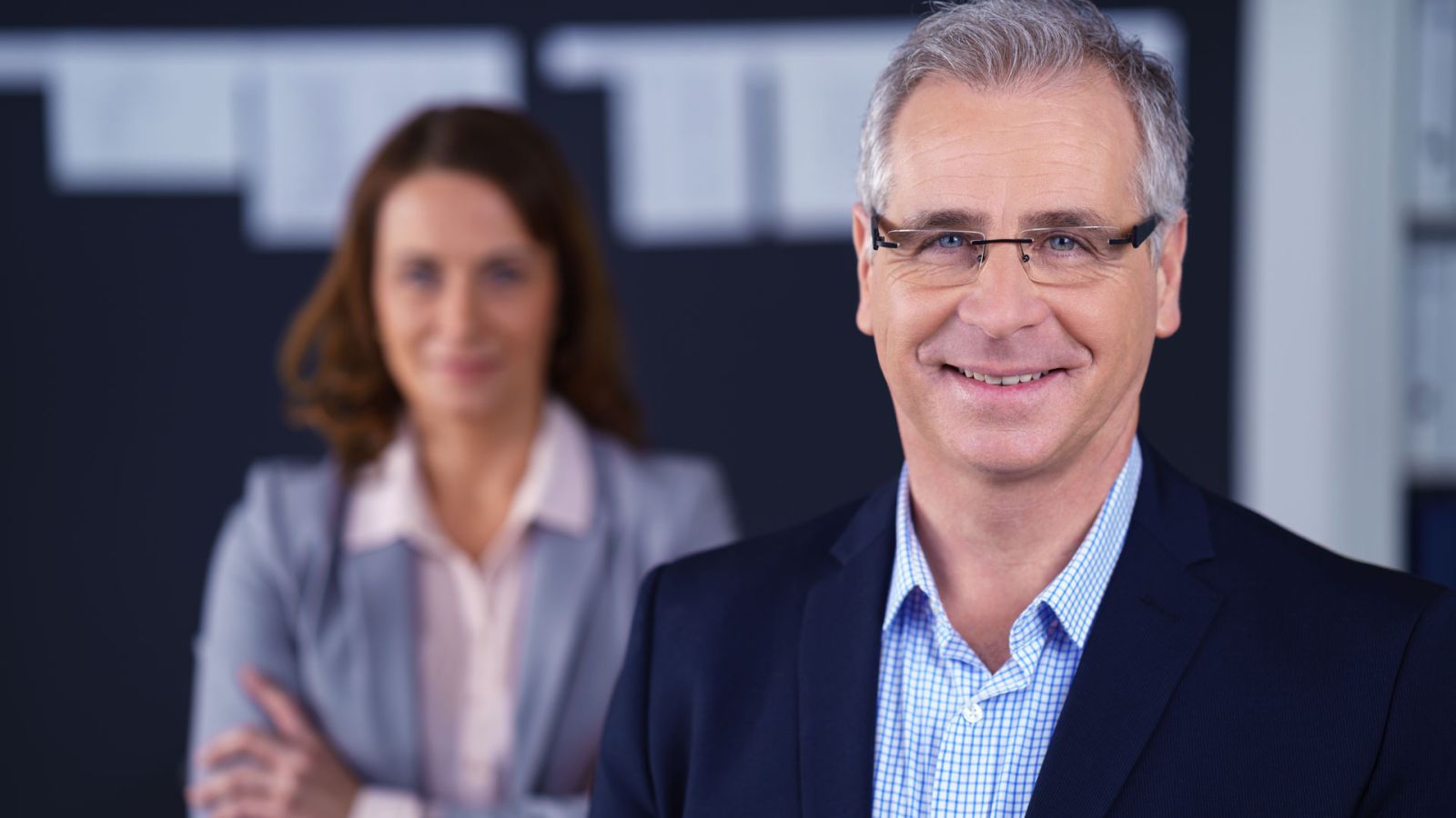 Porträt zweier Fachkräfte in Businesskleidung in einer modernen Büroumgebung: Ein lächelnder Manager im Vordergrund, eine Kollegin mit verschränkten Armen unscharf im Hintergrund. Ideal zur Darstellung von Leadership, Auditbereitschaft, Compliance und verlässlicher Qualitätssicherung. Porträt zweier Fachkräfte in Businesskleidung in einer modernen Büroumgebung: Ein lächelnder Manager im Vordergrund, eine Kollegin mit verschränkten Armen unscharf im Hintergrund. Ideal zur Darstellung von Leadership, Auditbereitschaft, Compliance und verlässlicher Qualitätssicherung.